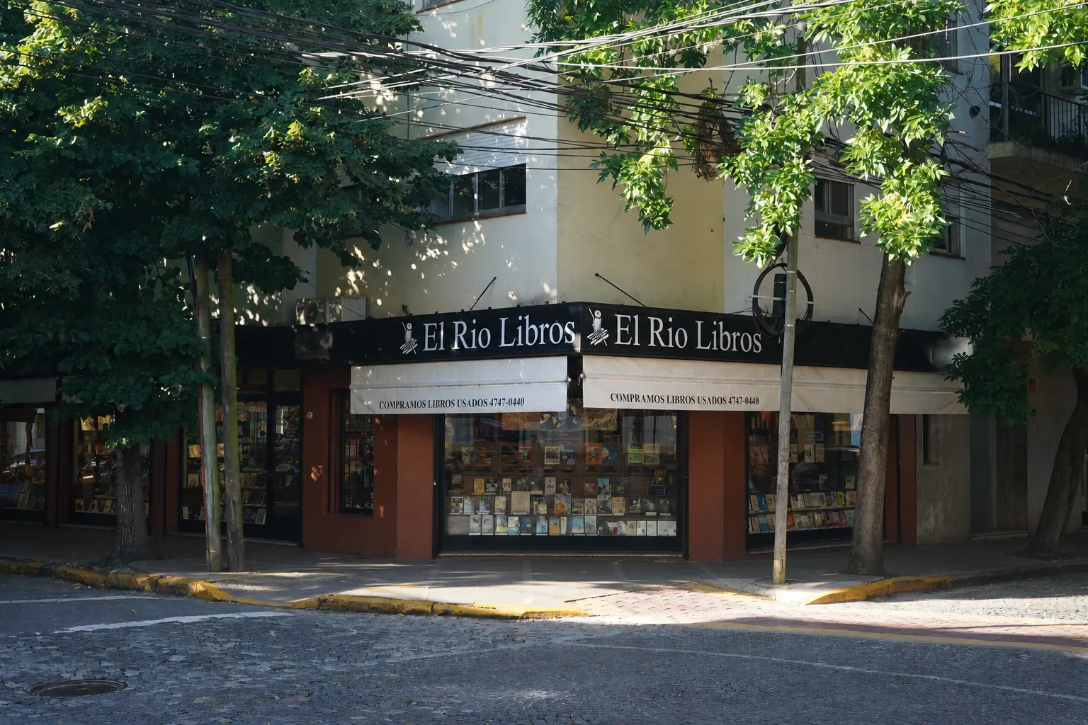 Frente de El Río Libros en la esquina de Acassuso 215, San Isidro: dos vidrieras llenas de libros bajo toldos claros que anuncian la compra de usados.